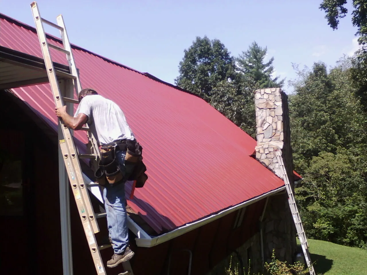 Roofer on a ladder beside a red metal roof and stone chimney — Soffit & Fascia Repair by Roofing Brothers in Hudgins
